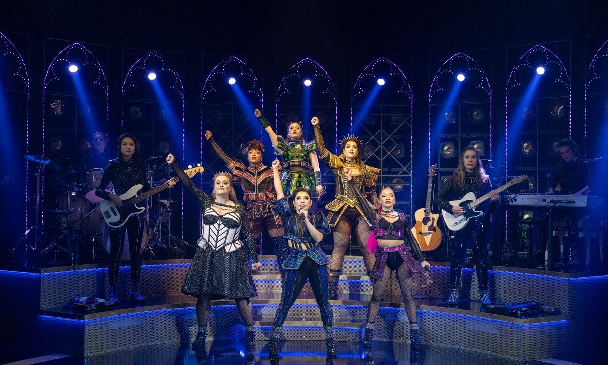 SIX the Musical cast raising fists in triumphant pose on stage with live all-female band behind them