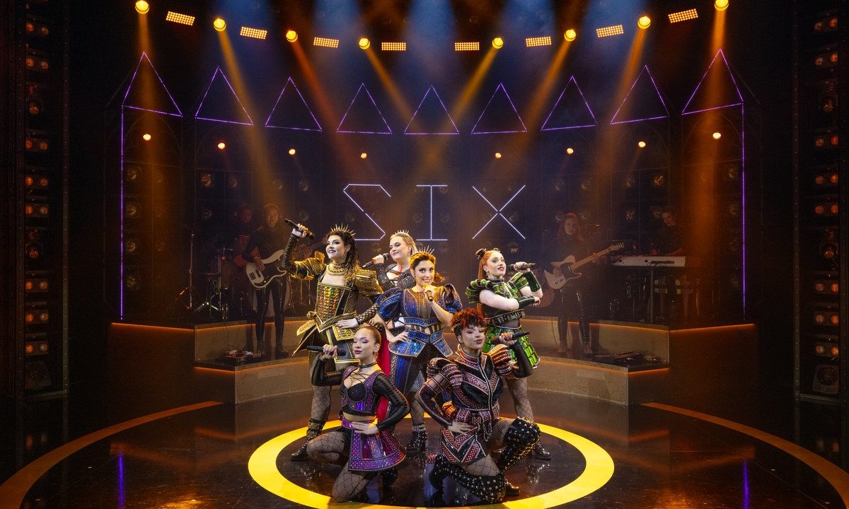 SIX the Musical cast posed beneath illuminated SIX sign with gold and purple stage lighting
