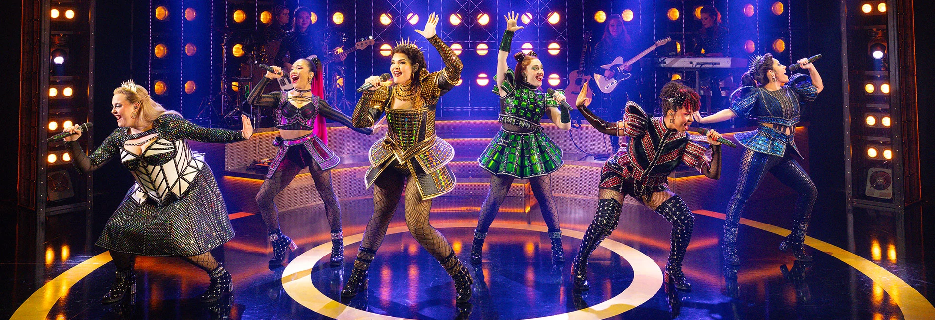 SIX the Musical cast performing on stage in jewelled Tudor-pop costumes under blue and gold stage lights