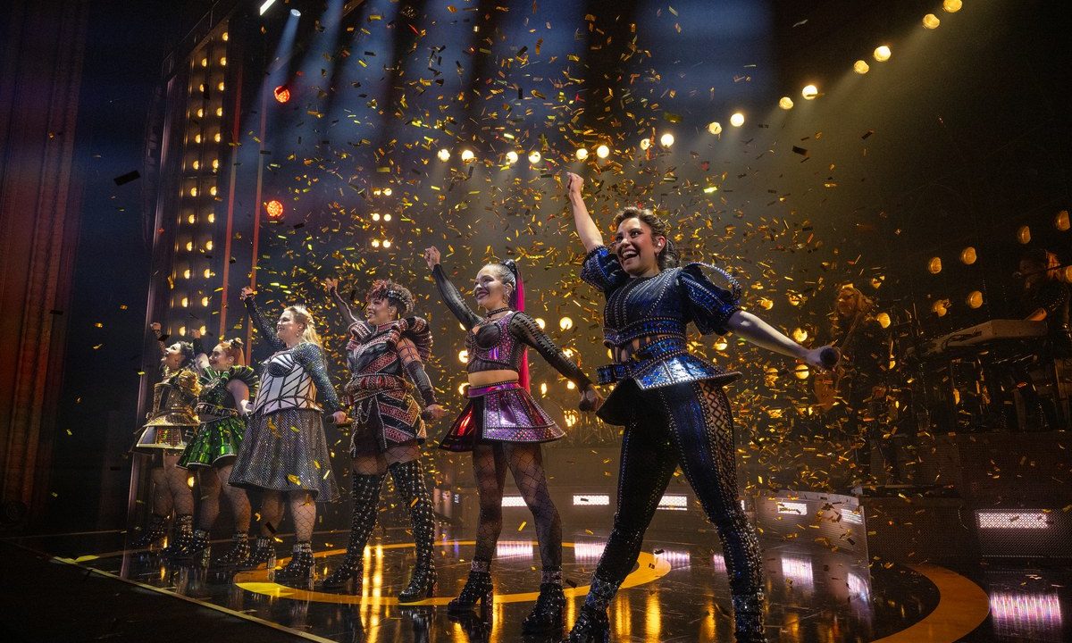 SIX the Musical cast celebrating with raised arms as gold confetti falls on stage